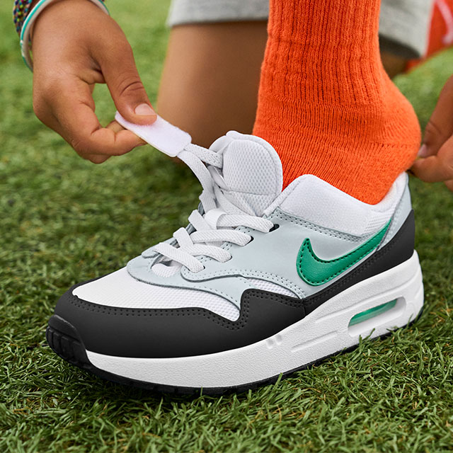 Person velcroing their Nike Easy On Air Max sneaker with a white, gray, and black upper, green swoosh, and visible air unit, while standing on grass in bright orange socks.
