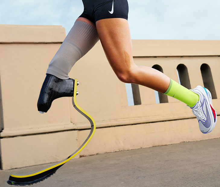 Person running with a black prosthetic blade on the left leg and a white Nike running shoe with red and blue accents on the right, wearing a neon green sock and black Nike shorts