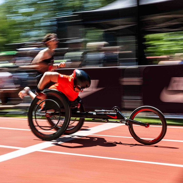 Nike employee competing in a one-mile race alongside runners, wearing an orange racing outfit and helmet, crossing the finish line in a racing wheelchair.